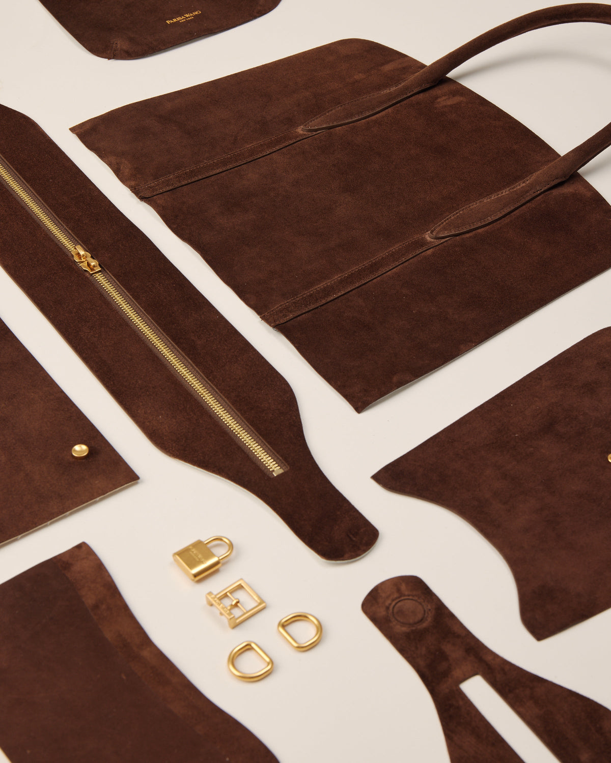 Chocolate suede components, handles, zipper, and hardware arranged before handbag assembly