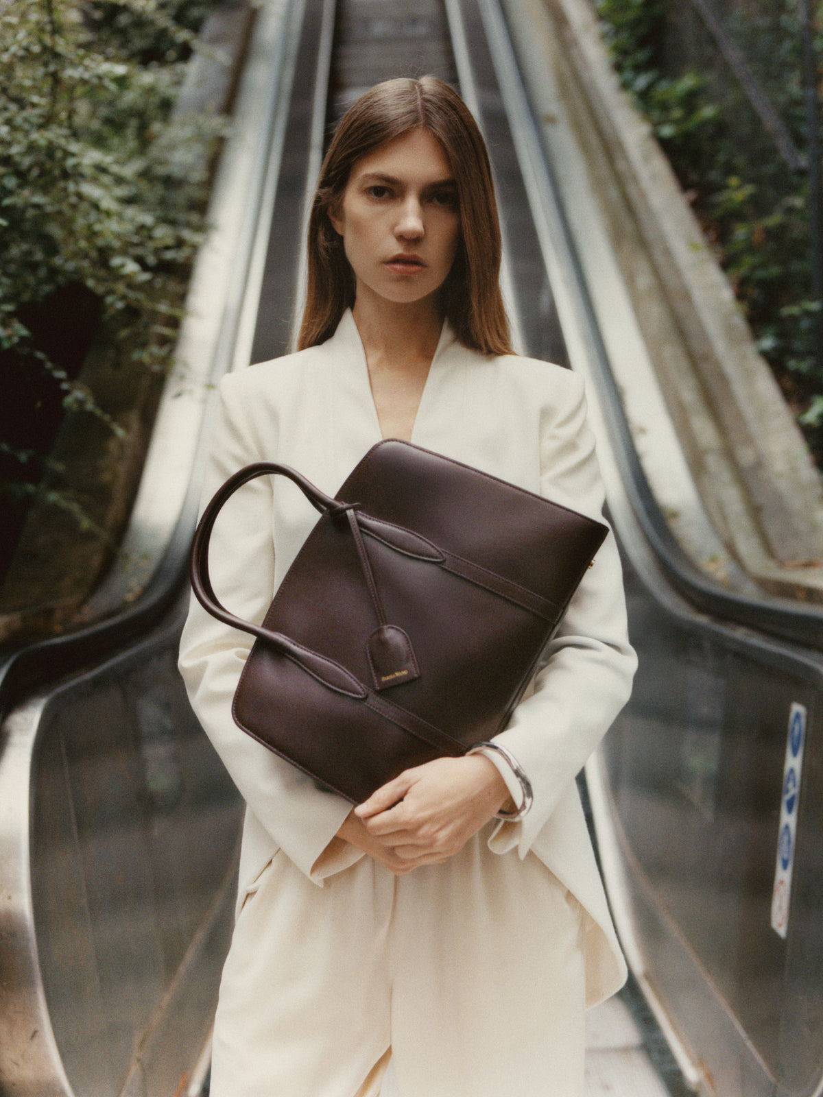 Summer 2026 campaign: model in white tailoring carrying a chocolate Gabrielle top-handle bag.