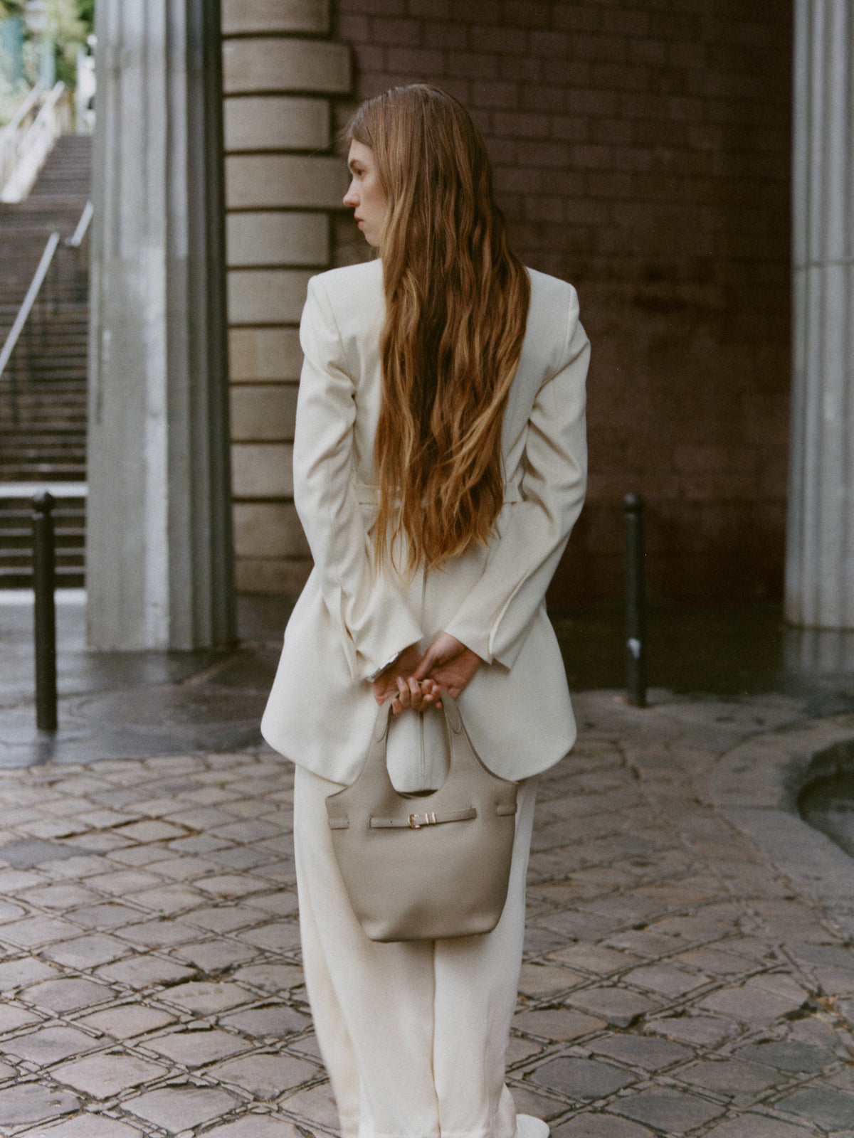 Summer 2026 campaign: model in white tailoring carrying a cream leather handbag.