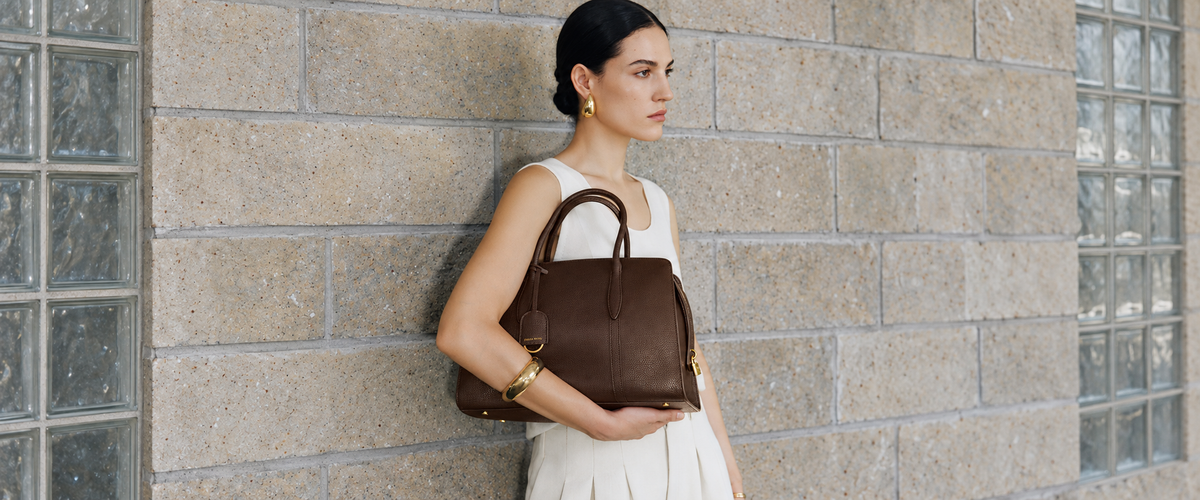 Fall campaign: model carrying a chocolate pebbled Gabrielle 35 against stone and glass-block architecture.