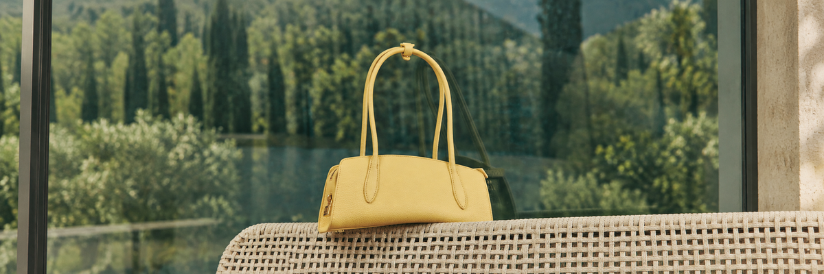 Campaign photograph: yellow pebbled East-West shoulder bag on woven rattan surface, green woodland/window backdrop; background extended for a landscape banner.