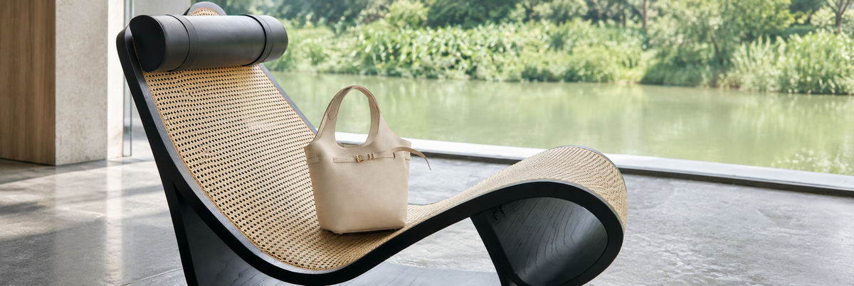 Campaign photograph: cream pebbled bucket handbag resting on sculptural woven cane chair, green garden view; background extended for a landscape banner.